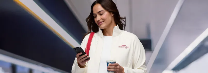 A woman in a branded white corporate uniform jacket using a smartphone while holding coffee.