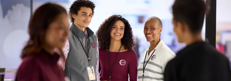 Three smiling employees wearing burgundy and gray branded uniforms with lanyards at a corporate event