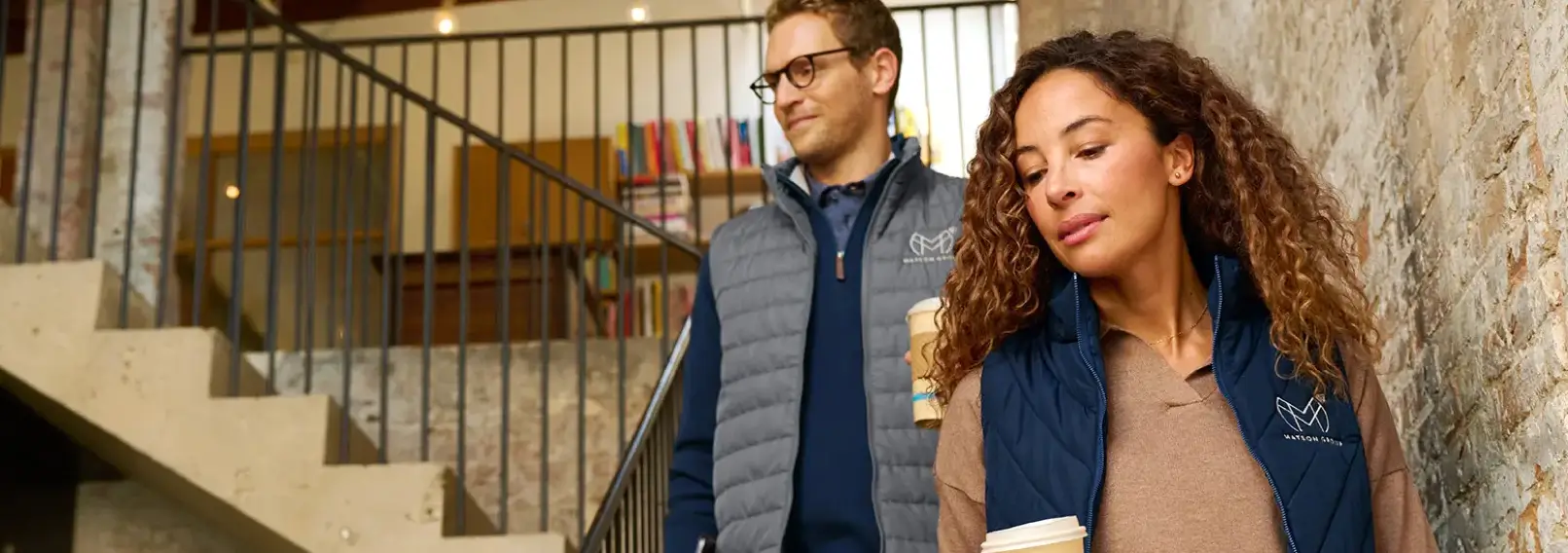 Two employees in quilted vests, descending office stairs while holding coffee