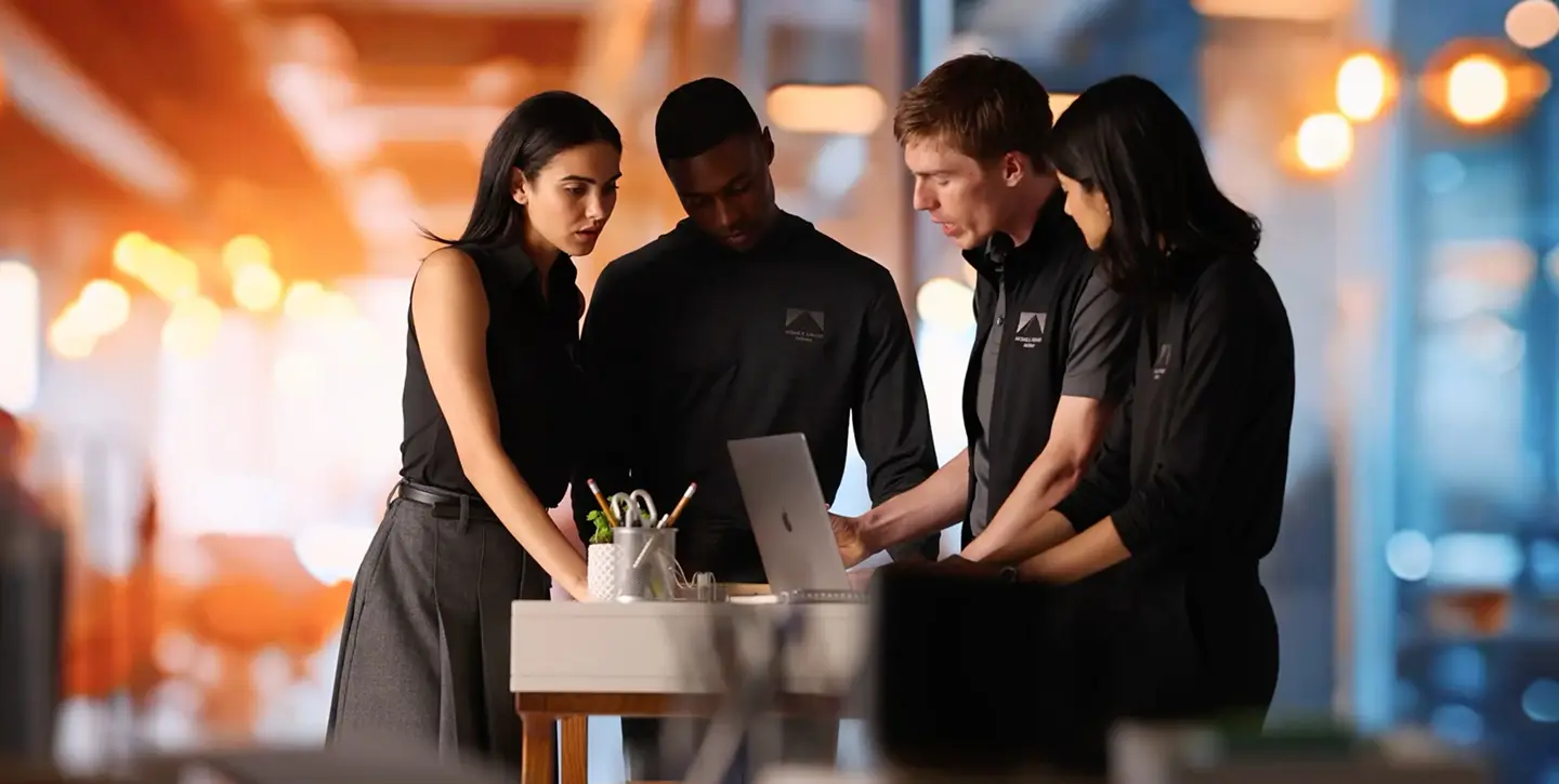 Four employees wearing matching branded apparel work around a laptop.