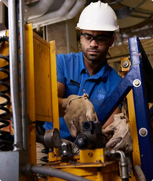 A man wearing a white hard hat and a blue polo works with machinery.