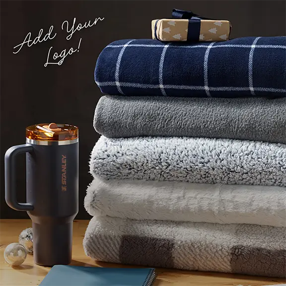 Add Your Logo! A stack of blankets of various patterns next to a drinkware mug.